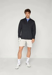Sudadera negra con cremallera sobre un polo azul claro, combinada con pantalones cortos claros y zapatillas deportivas blancas. El modelo sostiene un guante de golf.