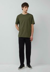 Olivegrünes kurzärmliges T-Shirt aus Baumwolle, mit Rundhalsausschnitt, kombiniert mit schwarzen Hosen und schwarzen Sneakers vor einem hellen Hintergrund.
