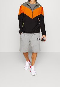 Man i svart, orange och grå zip-up jacka med grå shorts och vita sneakers, hållande en svart vattenflaska i ena handen.