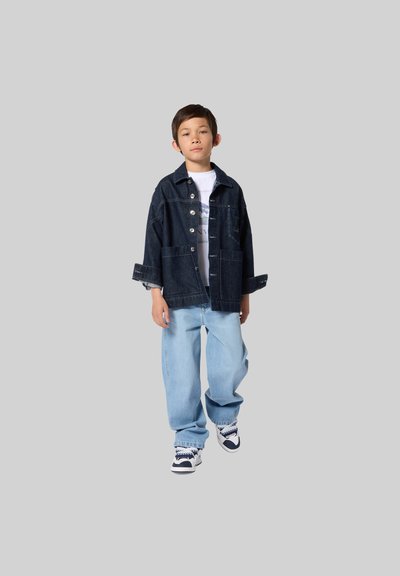 Donker denimjack met knopen, twee voorzakken en opgerolde mouwen, gecombineerd met lichtblauwe losse jeans en sneakers.