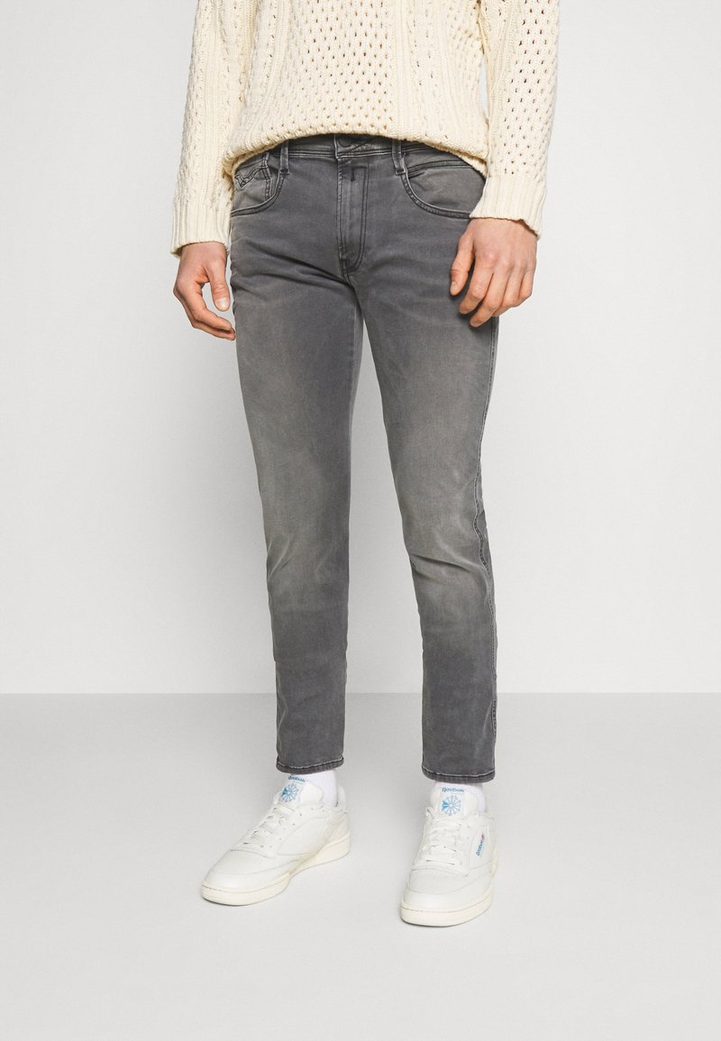 Jeans de mezclilla gris con corte slim, ligero desgaste, diseño de cinco bolsillos y detalles de costura, combinados con zapatillas blancas y calcetines de caña corta.