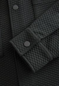 Prošívaná tmavě šedá látka se strukturovaným diagonálním vzorem, s černým patentním knoflíkem na manžetě a elegantním designem.