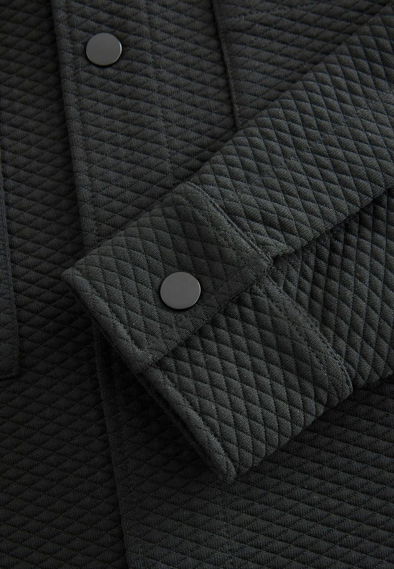 Tessuto trapuntato grigio scuro con motivo diagonale strutturato, caratterizzato da un bottone automatico nero sul polsino e un design lineare.