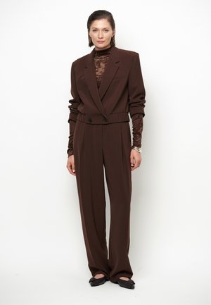 Frau trägt einen braunen cropped Blazer und weite Hosen über einem transparenten braunen Rollkragentop, stehend vor weißem Hintergrund.