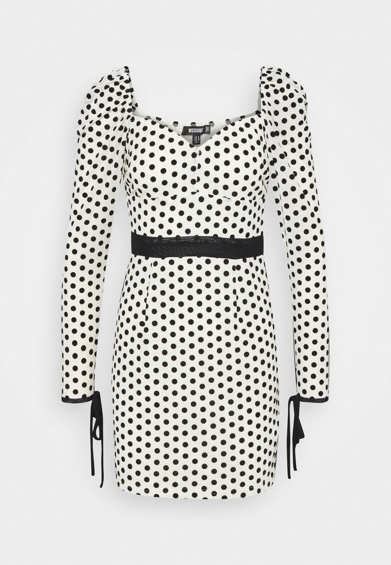 Robe mini blanche à manches longues et pois noirs, col carré, épaules bouffantes, ceinture noire à la taille et liens aux poignets des manches.