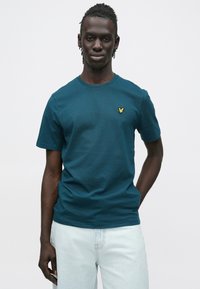 Νεαρός άνδρας με πλεξούδες, φοράει τιρκουάζ κοντομάνικο πουκάμισο με μικρό κίτρινο λογότυπο πουλιού και ανοιχτόχρωμα μπλε τζιν, στέκεται μπροστά σε λευκό φόντο.