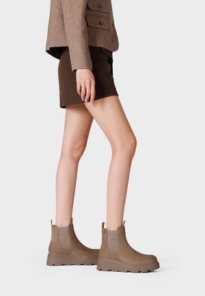 Donna che indossa stivali alla caviglia in pelle scamosciata marrone, pantaloncini marroni e un blazer color taupe, in piedi su sfondo bianco.