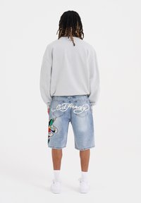 Felpa grigia oversize e pantaloni shorts in denim blu con un design grafico colorato e il testo "Ed Hardy" sulla schiena. Sneakers bianche completano il look.