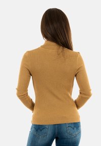 Maglione a collo alto a coste di un caldo colore beige, con maniche lunghe e una silhouette aderente, abbinato a jeans blu con dettagli sulle tasche posteriori.