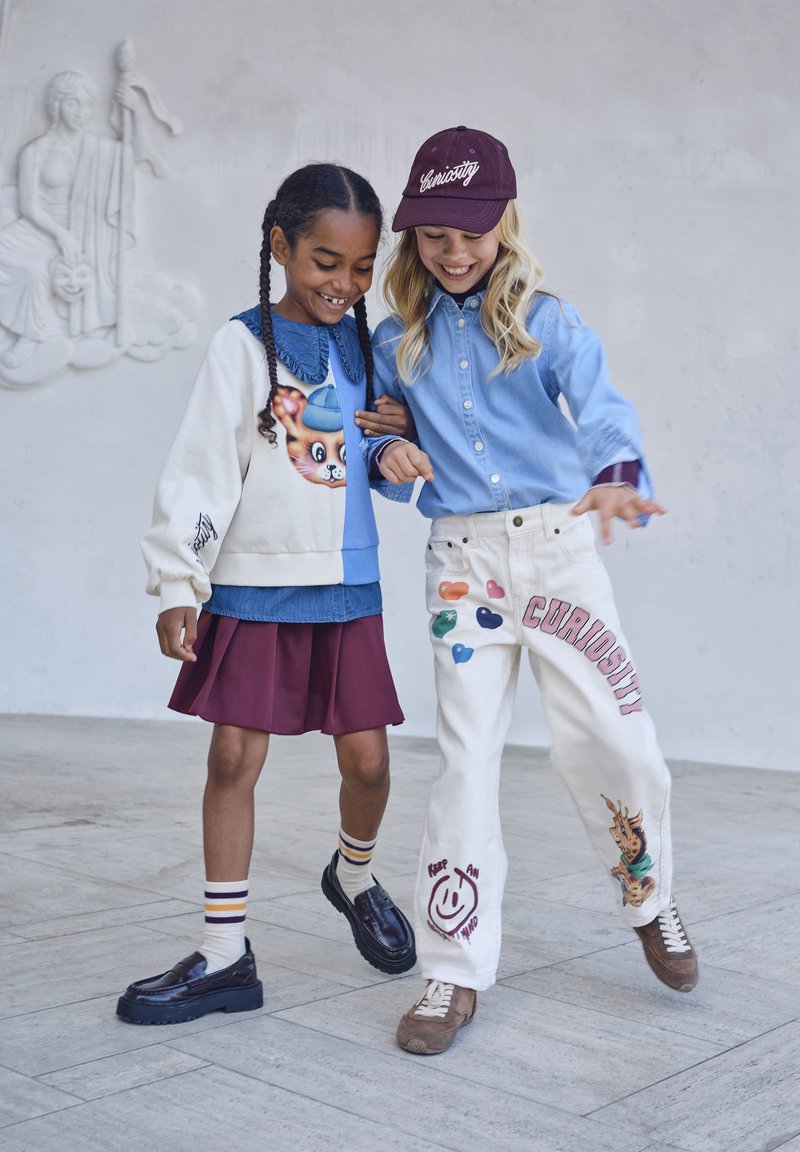 Deux filles souriantes marchent bras dessus bras dessous à l'intérieur ; l'une porte un sweat avec un chat et une jupe, l'autre porte une casquette « Curiosity » et un pantalon blanc décoré.