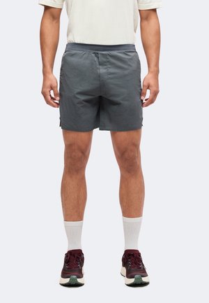 Mann trägt dunkelgraue Sportshorts, weiße Knöchelsocken und dunkelrote Laufschuhe und steht vor einem einfarbigen Hintergrund.