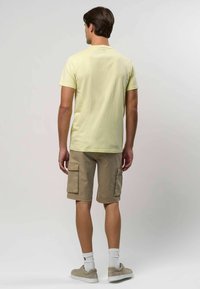 Svijetložuta pamučna majica, kaki cargo kratke hlače s dvostrukim bočnim džepovima, bijele čarape do gležnja i bež tenisice. Prikazan stražnji pogled.