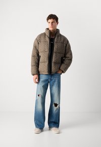 Polstret quilted jakke i tekstureret taupe med lynlås, parret med lyseblå slidte vide jeans og beige sko.