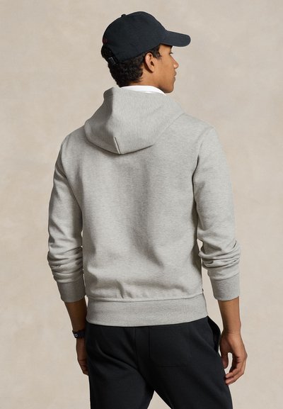 Mand iført lysegrå hættetrøje og sort kasket, vist bagfra mod en beige baggrund.