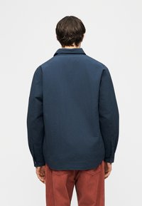 Άνδρας με σκούρα μαλλιά που φορά ένα μακρυμάνικο μπλε μπλουζάκι ναυτικού τύπου και παντελόνι σε χρώμα σκουριάς, όρθιος με το πρόσωπο στραμμένο προς το αντίθετο μέρος, μπροστά σε ένα απλό φόντο.