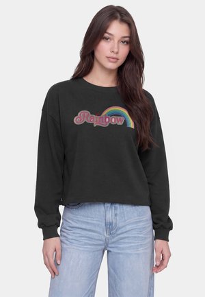 Giovane donna con lunghi capelli castani che indossa una felpa nera con il testo "Rainbow" e una grafica arcobaleno, abbinata a jeans azzurri chiari.
