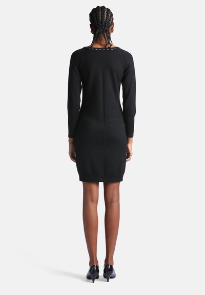 Femme aux cheveux tressés portant une robe noire moulante à manches longues et des chaussures noires à talons, debout de dos sur un fond blanc.