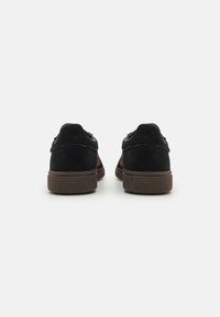 Baskets en suede noir avec une semelle en caoutchouc marron texturé, présentant un branding minimal et un design simple et classique sans détails complexes.