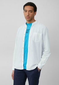 Witte button-up shirt met een zachte textuur, voorzien van een zak, gedragen over een felblauw t-shirt met zwarte tekst. Geen zichtbare patronen.