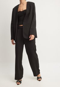 Melnas pielāgotas žaketes ar iegrieztu apkakli, apvienota ar melnu topu bez piedurknēm un atbilstošiem platajiem biksēm, valkāta ar melnām atvērtā purngala kurpēm.