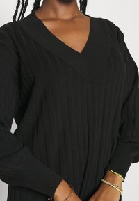 Pull noir en tricot côtelé avec un col en V, manches longues et une finition texturée. Accents avec des bracelets superposés au poignet.