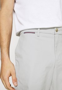 Pantalones gris claro hechos de un tejido suave, con un corte ajustado, bolsillos laterales y un sutil acento de rayas cerca de la cinturilla. Detalle del botón dorado visible.