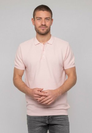 Homme avec cheveux courts et barbe portant un polo à manches courtes rose clair et un jean gris, debout les mains jointes devant lui.