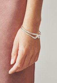 Bracciale d'argento con superficie liscia e lucida, caratterizzato da un design unico ad attorcigliamento e un piccolo diamante come accentuazione alla chiusura.