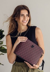 Bordeauxfarbene Tasche mit Muster und glatter Textur, ausgestattet mit einem Reißverschluss und schwarzen Akzenten, gehalten von einer Person in einem schwarzen Oberteil.