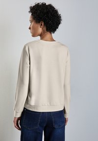 Ljusbeige sweatshirt med rund halsringning och ribbade ärmar, med en avslappnad passform och mjuk textur, kombinerad med mörka denimjeans.
