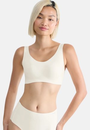 Giovane donna con capelli biondi corti che indossa un reggiseno sportivo bianco senza cuciture e slip a vita alta abbinati, su sfondo semplice.