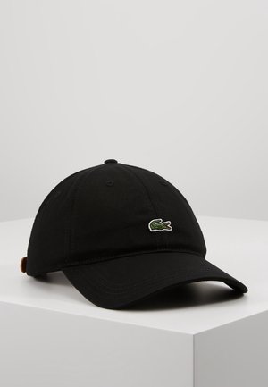 Gorra de béisbol negra con visera curva, logo de cocodrilo verde en el frente y correa ajustable marrón en la parte trasera, colocada sobre una superficie blanca.
