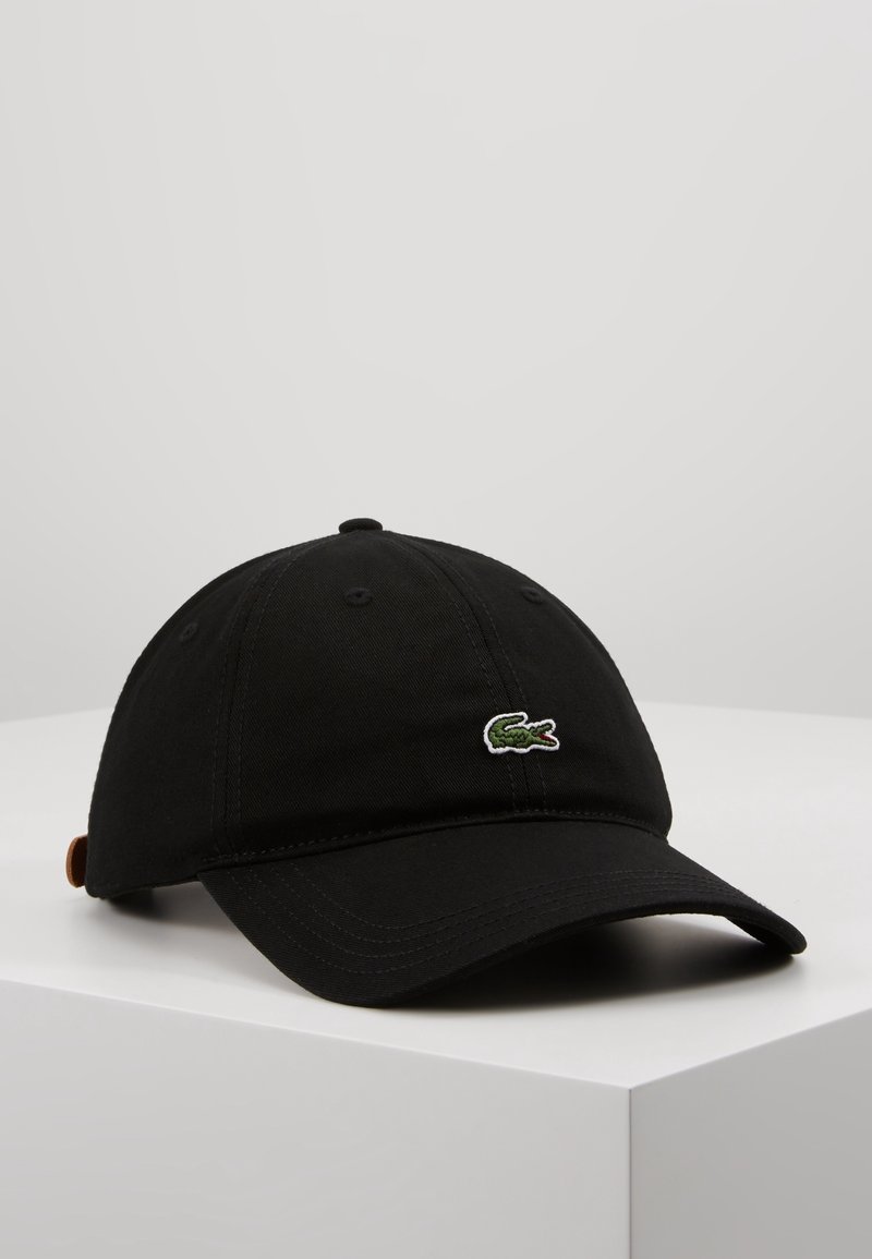 Casquette de baseball noire avec une visière courbée, un logo crocodile vert à l'avant et une sangle marron réglable à l'arrière, placée sur une surface blanche.