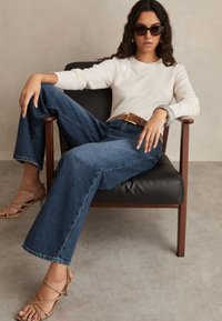 Vrouw met krullend haar, zonnebril, witte trui, wijde jeans en beige hakken, zittend in een zwarte leren stoel met houten armleuningen.