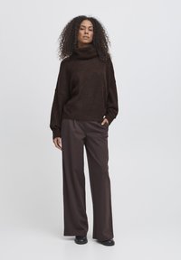 Maglione marrone oversize con collo a pompa in tessuto testurizzato, abbinato a pantaloni marroni a vita alta e gamba larga e stivaletti neri alla caviglia.