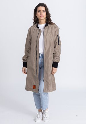 Khaki long bomber jacket with zip closure, ribbed cuffs, side pockets, and an orange label, paired with light blue jeans and white sneakers.