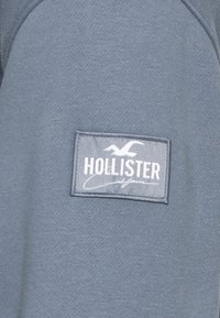 Grå sweatshirt med tygstruktur och ett fastsytt lädermärke med texten "Hollister California" och en fågel-logotyp.
