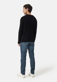 Svart stickad tröja med texturerad yta, parat med blå denimjeans och vita sneakers, sedd bakifrån.