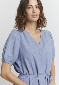 Světle modré šaty s krátkým rukávem a výstřihem do V, s nařasenými puff rukávy a páskem v pase. Látka má texturovaný povrch s jemnými variacemi.