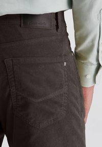 Nahaufnahme einer Person, die dunkelbraune Cordhosen mit einer Gesäßtasche und einem hellgrünen, langärmeligen Hemd trägt, das an den Handgelenken hochgekrempelt ist.