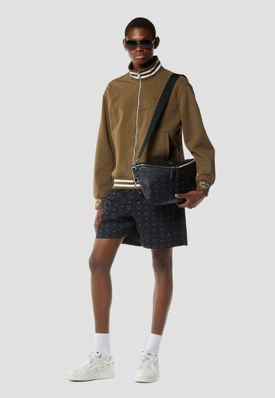 Man draagt een bruin jack met ritssluiting, zwarte korte broek met patroon, witte sneakers, zonnebril en houdt een zwarte schoudertas vast tegen een effen achtergrond.