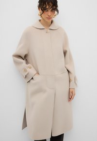 Cappotto in lana beige con colletto ampio, chiusura frontale a bottoni e tasche laterali. Presenta una vestibilità rilassata e polsini con bottoni, con una consistenza liscia.