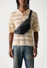 Schwarze Umhängetasche mit geometrischem Design und Reißverschluss, getragen über einem beigefarbenen Strickpolo mit Lochmuster und hellblauen Jeans.