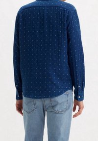 Camicia azzurra a maniche lunghe con una sottile texture e piccoli punti a motivo, caratterizzata da un orlo curvo e polsini con bottoni, abbinata a jeans blu chiaro.