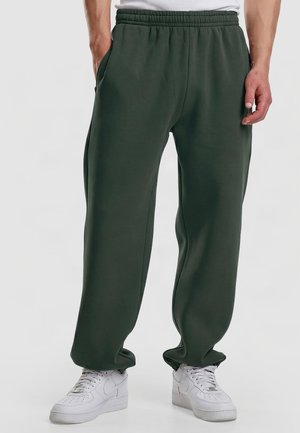 Homme portant un pantalon de jogging ample vert foncé à taille élastique et des baskets blanches, debout devant un fond clair uni.