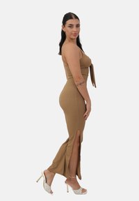Vestido maxi marrón de canalé con aberturas laterales, que presenta un diseño ajustado y un lazo ajustable en el busto. Estilizado con sandalias de tacón transparente.