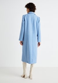 Casaco de lã azul claro com um design ajustado, mangas longas e uma costura central nas costas. Textura suave, comprimento até os joelhos, combinado com botas de cano alto em creme.