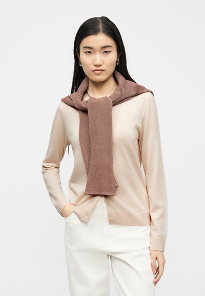 Νεαρή γυναίκα με μακριά μαύρα μαλλιά που φοράει μπεζ ζακέτα, λευκό παντελόνι και καφέ πουλόβερ περασμένο στους ώμους της, σε λευκό φόντο.