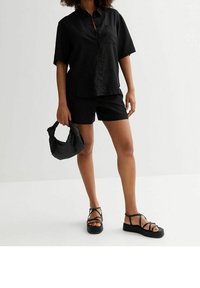 Chemise en lin noire à manches courtes, associée à un short noir. Le mannequin tient un sac noir texturé et porte des sandales noires à lanières.