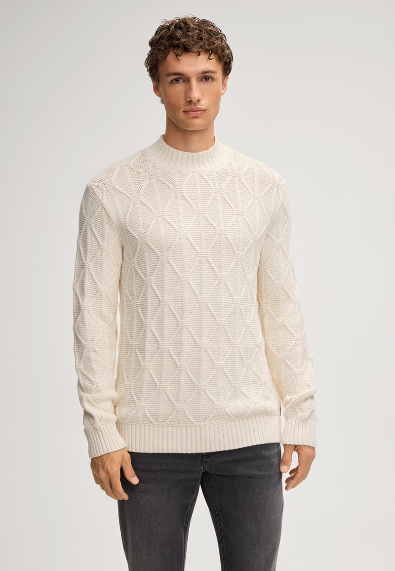 Beige-weißer Strickpullover mit geometrischem Muster, geripptem Kragen, Bündchen und Saum, kombiniert mit dunkelgrauen Jeans.